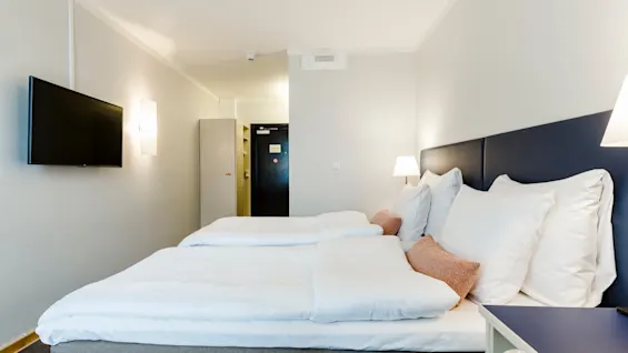 A neatly made double bed with white linens sits beside a nightstand and lamp, in a bright hotel room featuring a wall-mounted TV and a doorway in the background.