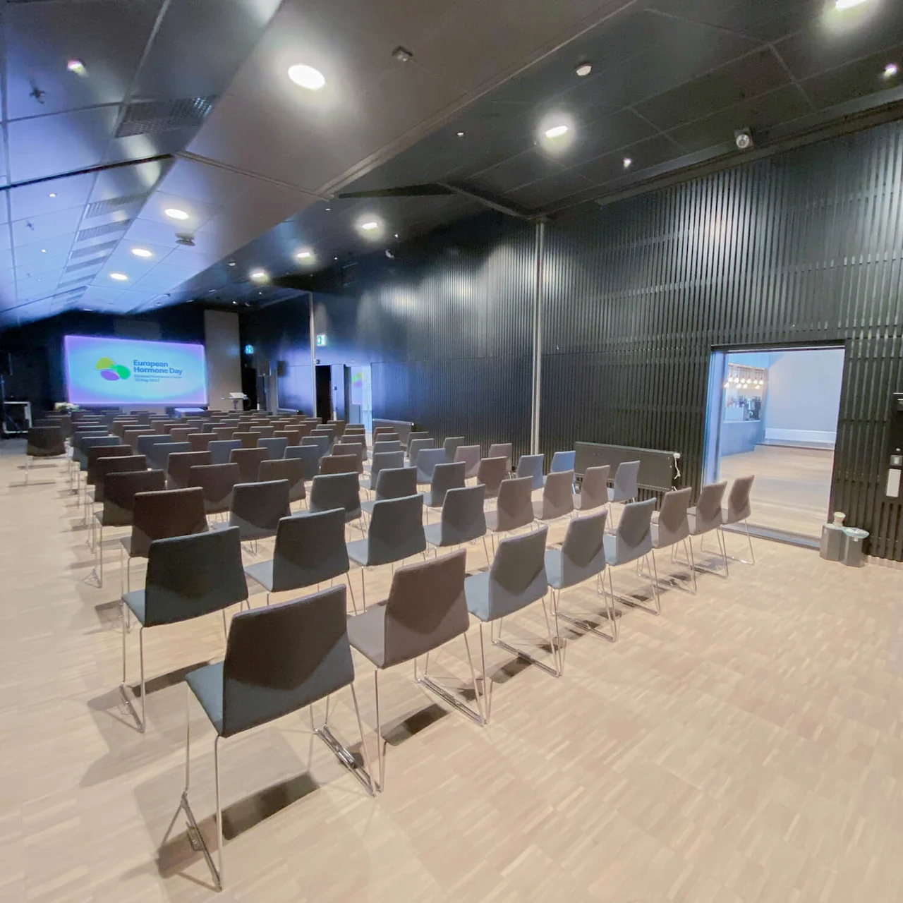 Norwegian Bokmål: En konferansesal med rader av tomme stoler vendt mot en skjerm som viser "European Hardware Awards." Rommet har moderne design, tregulv og svarte vegger.