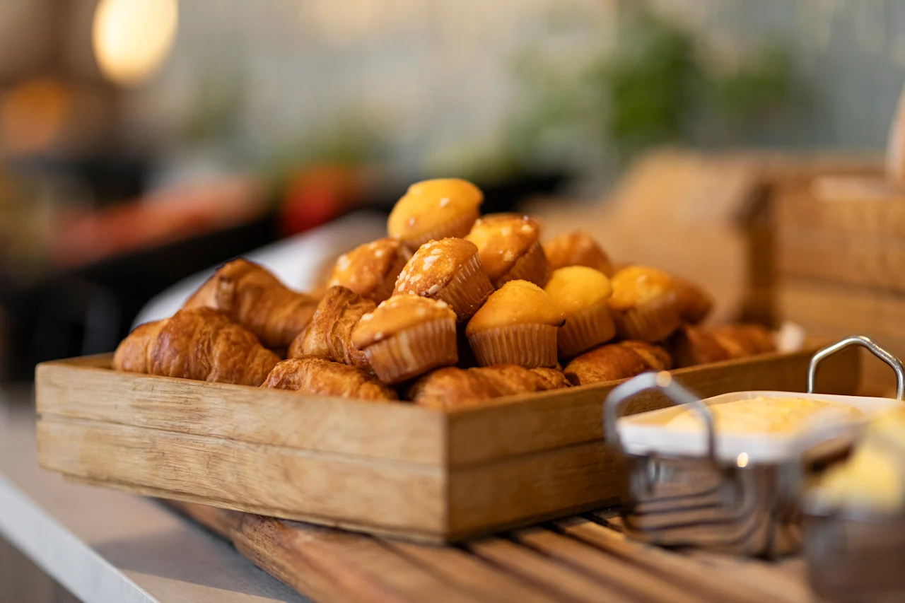 A wooden tray filled with golden-brown croissants and small muffins, ready to be enjoyed at a breakfast buffet.