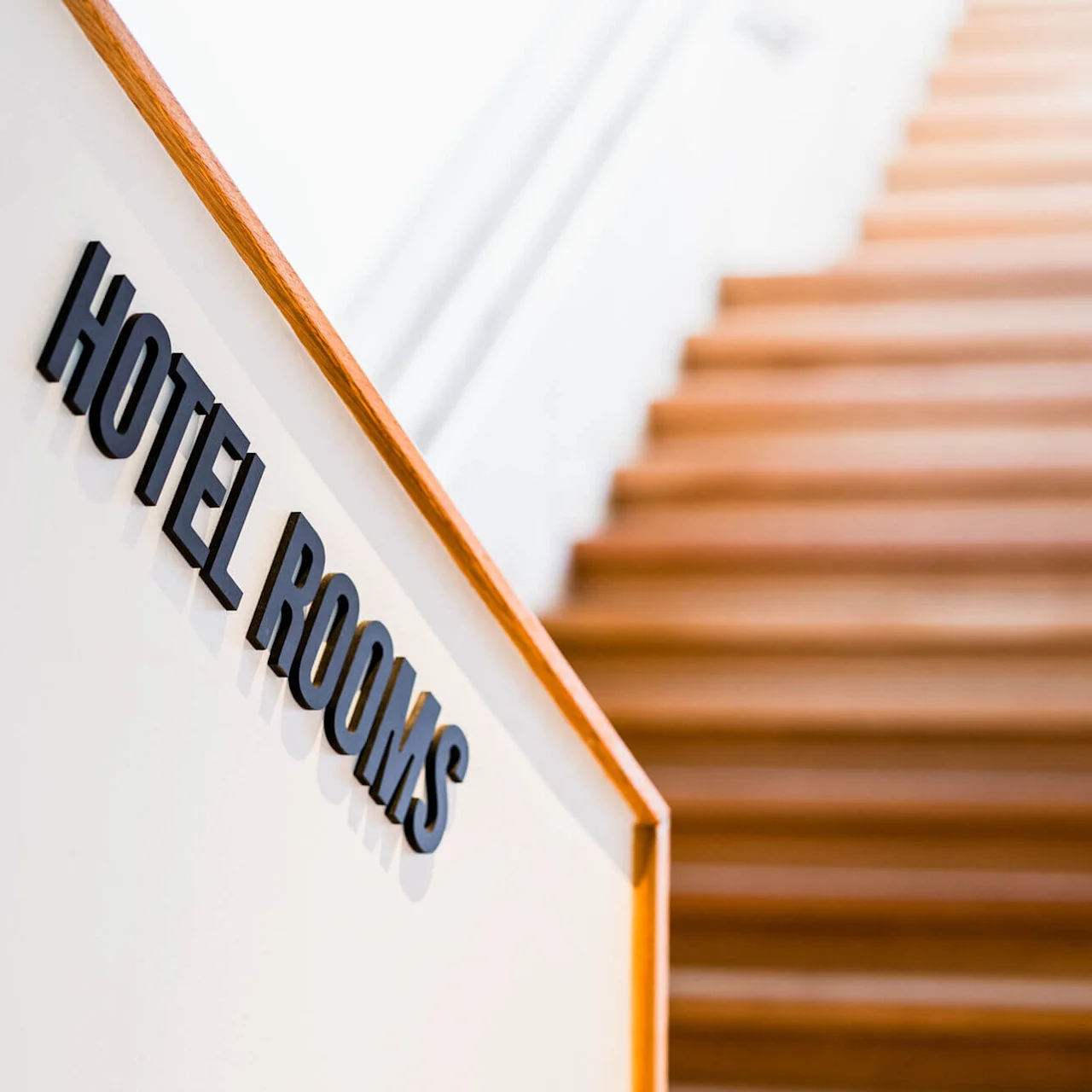 En skylt med texten "HOTEL ROOMS" är monterad på en vit vägg bredvid en trätrappa som går uppåt i en ljust belyst korridor.
