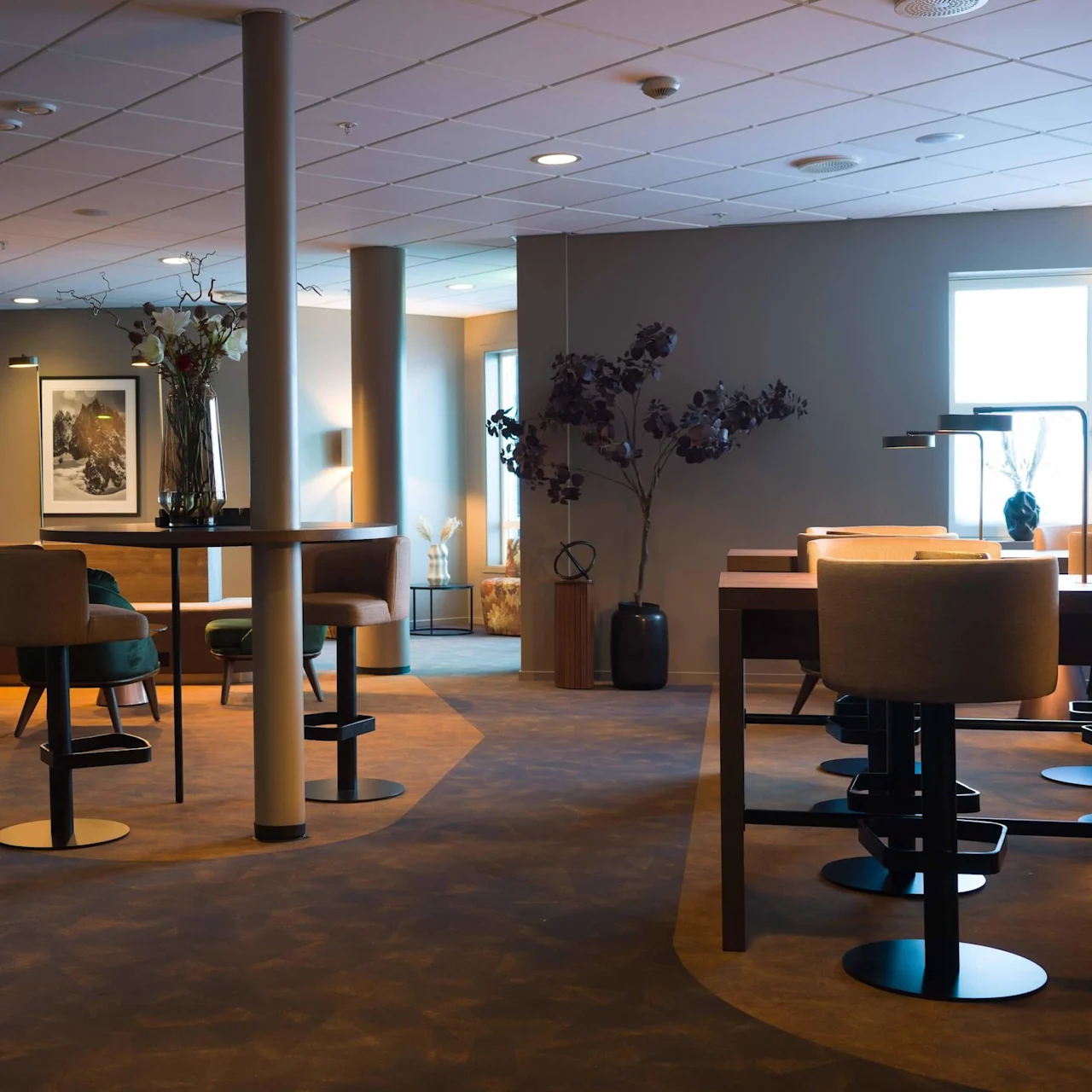 Chairs and tables are arranged in a cozy, modern lounge with soft lighting. Decorative plants and framed artwork adorn the space, creating a warm, inviting atmosphere. No text is visible.