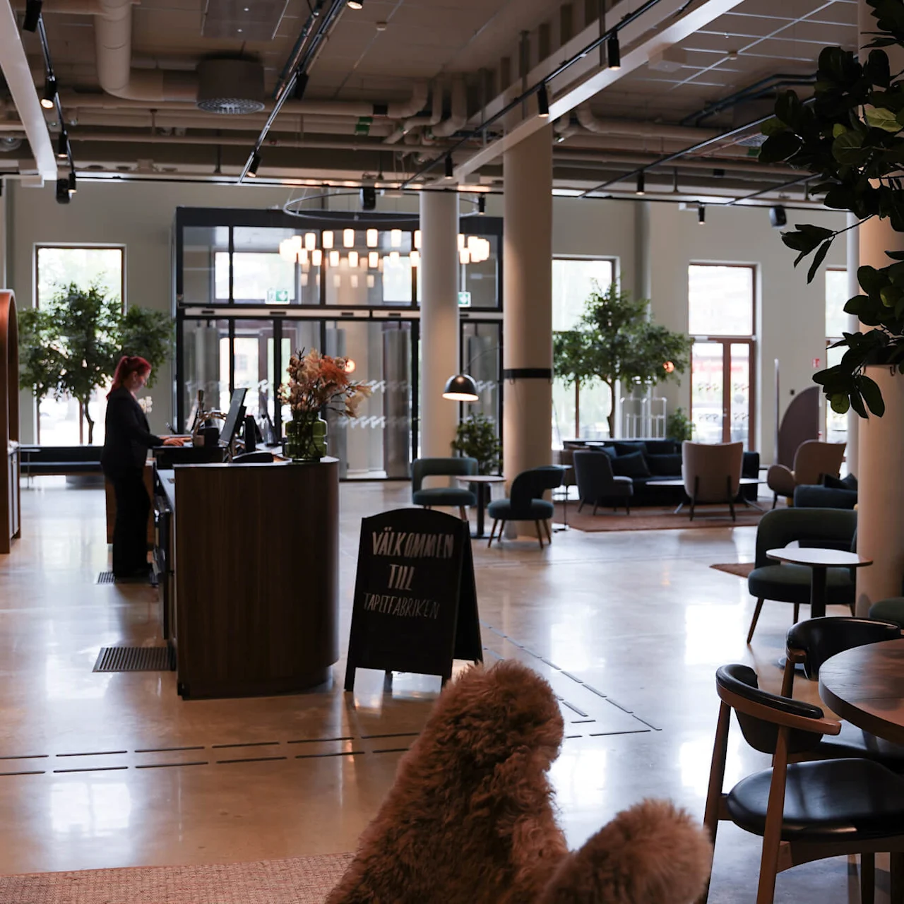 A person works at a sleek hotel reception desk amidst stylish lounge seating in an open, well-lit lobby.