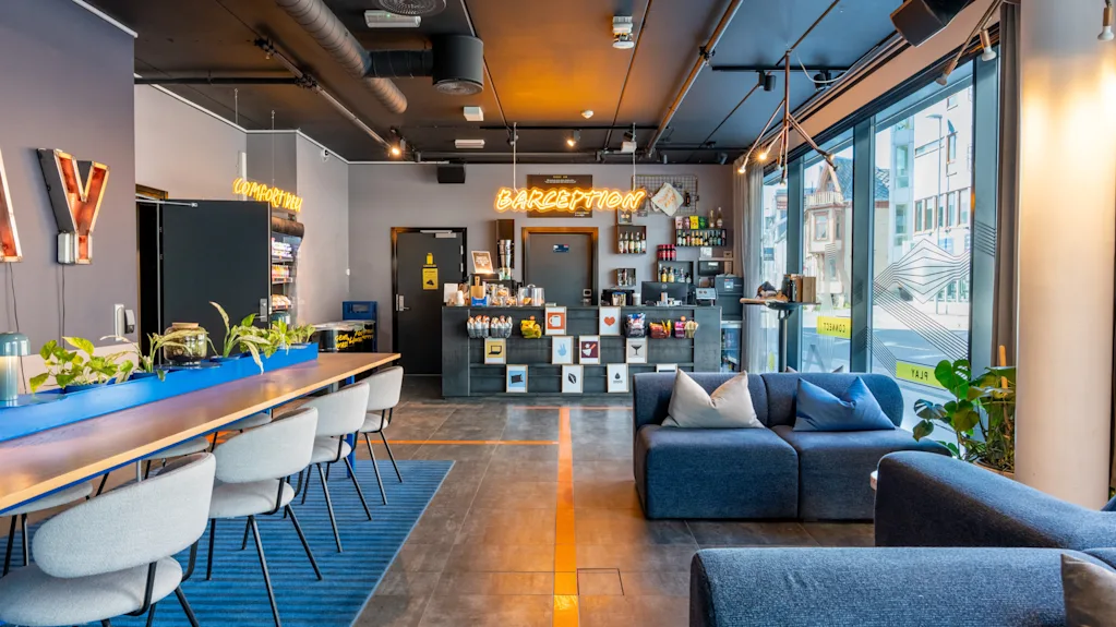 Moderni hotellin aula tyylikkäillä kalusteilla ja sisustuksella. Vastaanottotiski, jossa neonkyltti "CAFEPTION", ympärillään kaupunkimaisema suurien ikkunoiden läpi, mukavat istuimet ja modernit riippuvalaisimet.