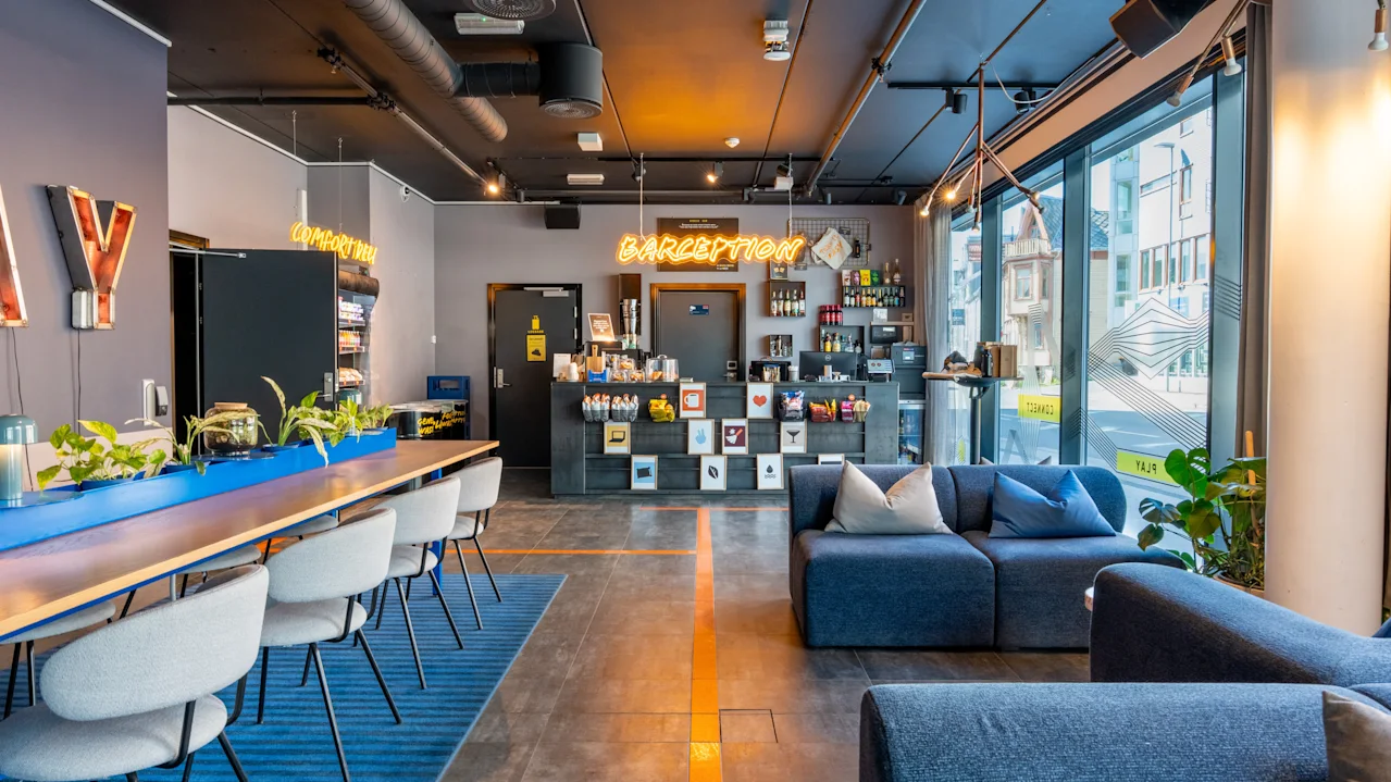 Moderni hotellin aula tyylikkäillä kalusteilla ja sisustuksella. Vastaanottotiski, jossa neonkyltti "CAFEPTION", ympärillään kaupunkimaisema suurien ikkunoiden läpi, mukavat istuimet ja modernit riippuvalaisimet.