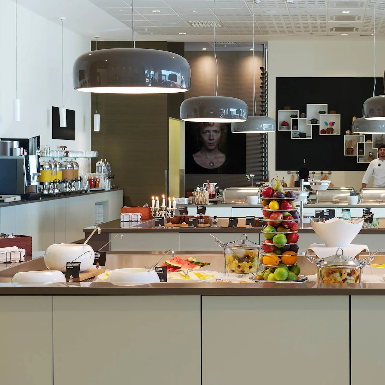 A buffet table is laden with various fruits and dishes; a chef stands behind. Modern pendant lights illuminate the contemporary dining area. Assorted juice dispensers and artworks decorate the space.