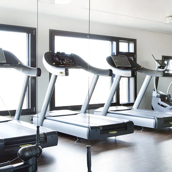 Treadmills and exercise equipment sit idle on a dark wooden floor in a bright, spacious gym with large windows and wall mirrors.
