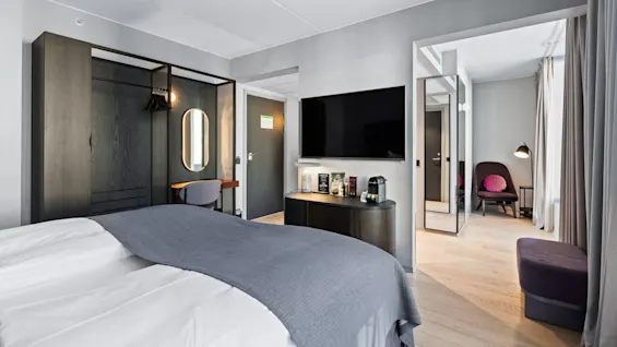 A modern hotel room features a neatly made bed with gray bedding, a desk and chair by a mirror, and a TV mounted on the wall. Adjacent area includes seating with a lamp.