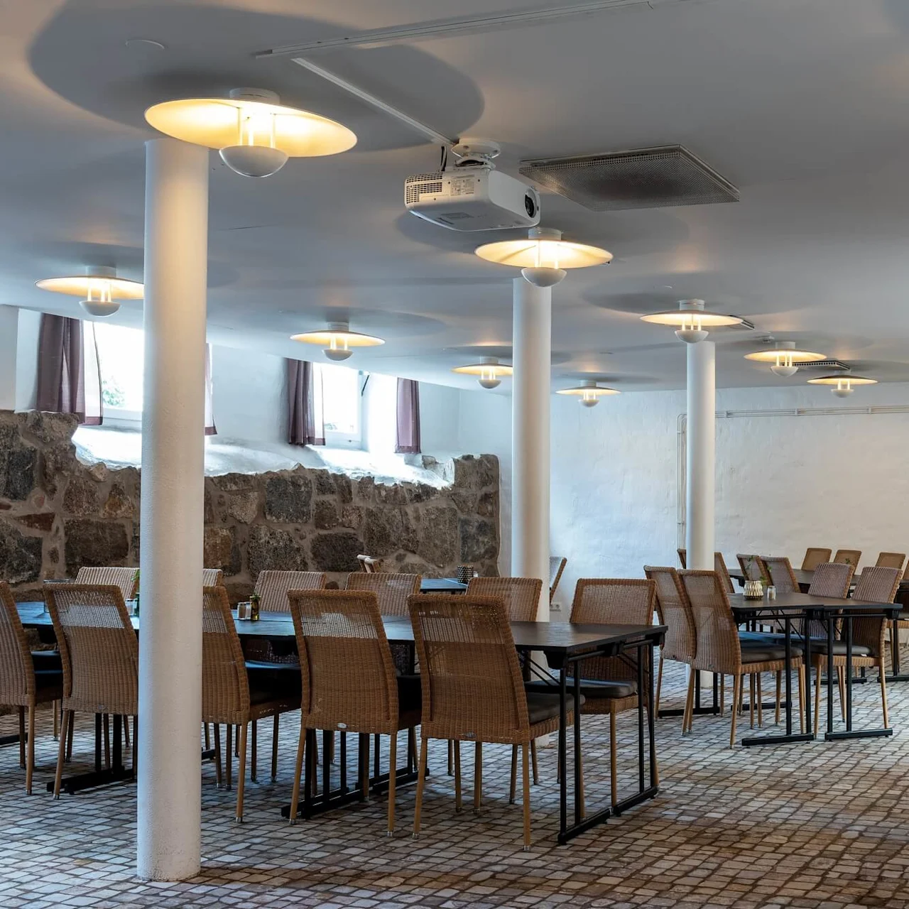 Ett tomt restauranginteriör med bord och stolar, golv med kullerstensmönster, vita pelare och moderna hängande lampor.