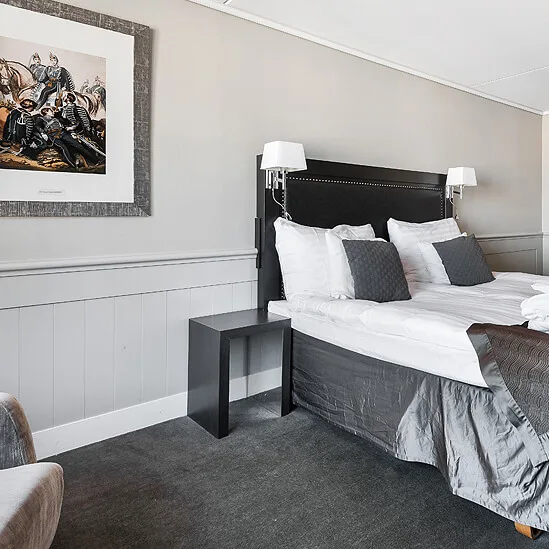 Neatly made bed with grey and white bedding in a tidy, modern bedroom setting with a wall-mounted artwork and dark carpet.