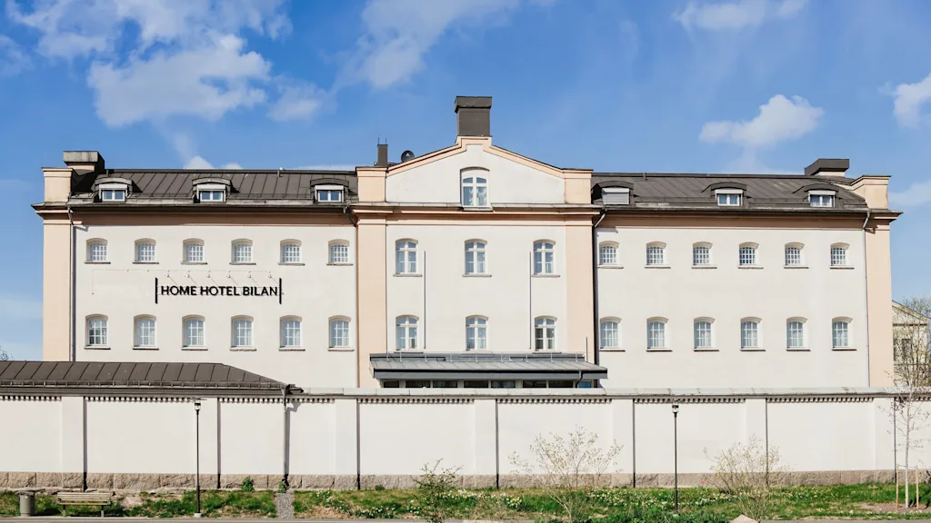 A large, light-colored building with many windows and a dark roof under a blue sky. A white wall stands in front of it. Text on building: HOME HOTEL BILAN.