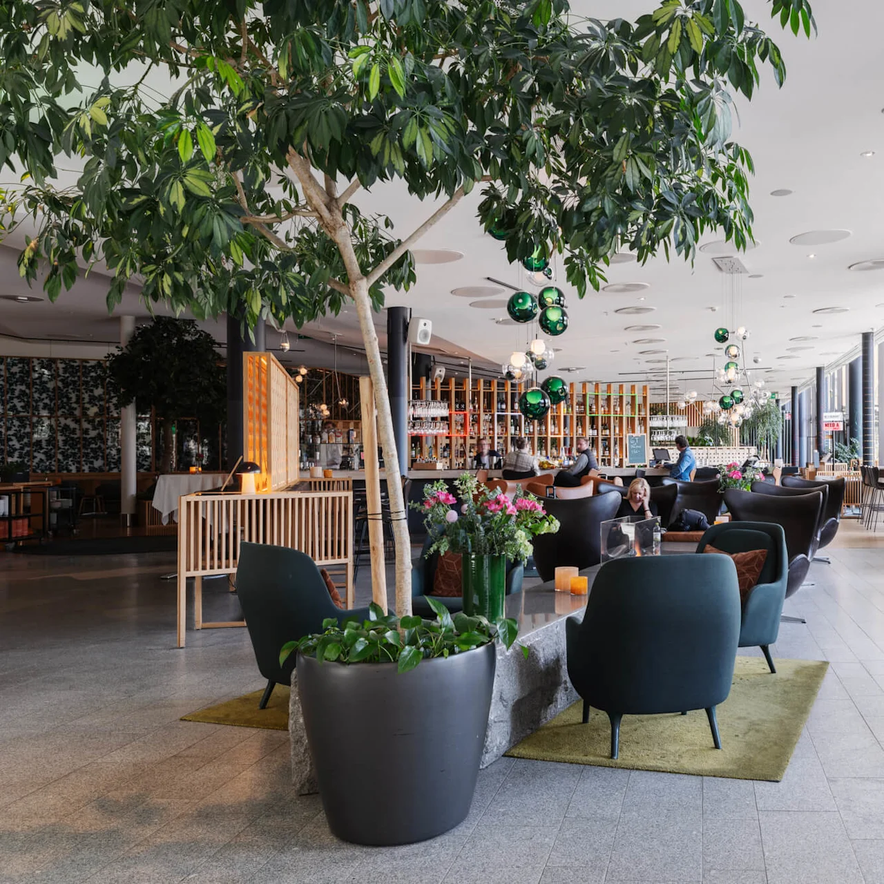 En mysig, modern lounge där människor sitter på mörka stolar vid bord, omgivna av stora växter, färgglada blommor och en bar. Skylten lyder "The Social Market."