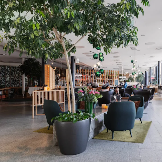 En mysig, modern lounge där människor sitter på mörka stolar vid bord, omgivna av stora växter, färgglada blommor och en bar. Skylten lyder "The Social Market."