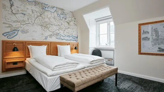 Et lyst og indbydende hotelværelse med to enkeltsenge, en stor kortvægskunst og et behageligt vinduessiddeområde.