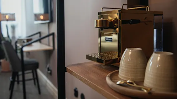 A golden coffee machine labeled "SJÖSTRAND" rests on a wooden shelf beside two cups, within a modern room featuring a television and seating area.