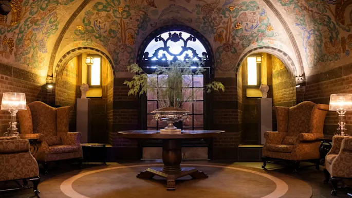 Elegant round table holding a decorative centerpiece; flanked by vintage armchairs in a vaulted room with intricate ceiling art and warm lighting from wall sconces, creating a cozy, luxurious atmosphere.