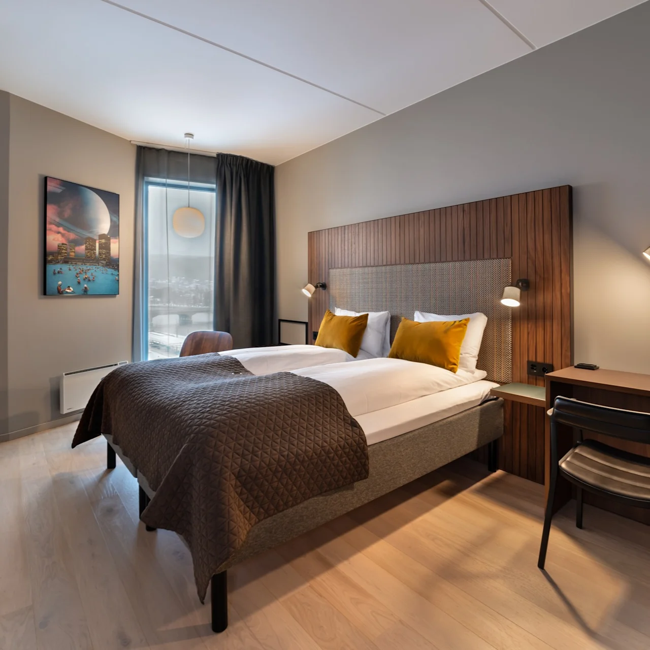 Et pent arrangert hotellrom med dobbelseng og gule puter, en trehodegjerde, et maleri på veggen, og et vindu med gardiner. Et skrivebord med lampe står i nærheten.