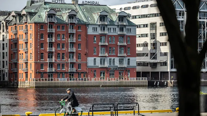 Clarion Hotel Admiral, a red brick building with balconies, overlooks a waterfront. "RESTAURANT" sign on facade. A person cycles by the riverside in the foreground.