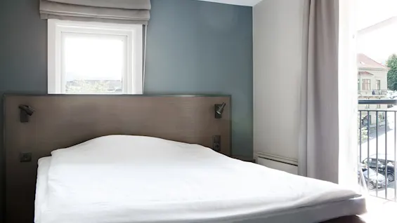 En pænt redt seng med hvidt sengetøj står i et moderne rum med blå vægge og et stort vindue, delvist dækket af gardiner, med udsigt til en gade udenfor.
