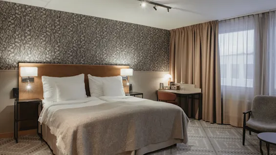 Ett mysigt hotellrum med en stor säng, mönstrad tapet, en skrivbordsplats och en bekväm fåtölj vid fönstret.