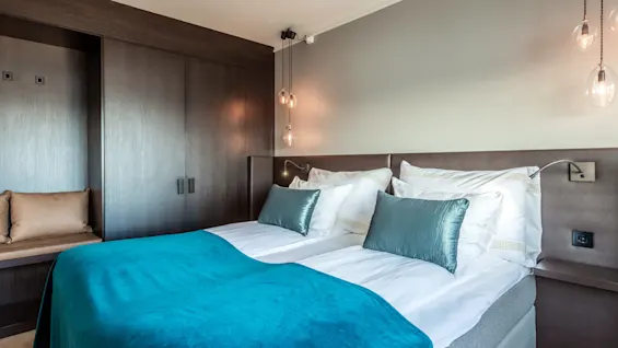 En pent oppredd seng med turkise detaljer står mot en mørk treveggsenhet med hengende lamper og en liten polstret benk. Rommet har en moderne, ren design.