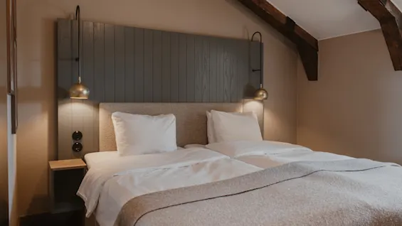 A cozy hotel room with two beds, a dark wooden headboard, and warm lighting from bedside lamps. Exposed beams add character.