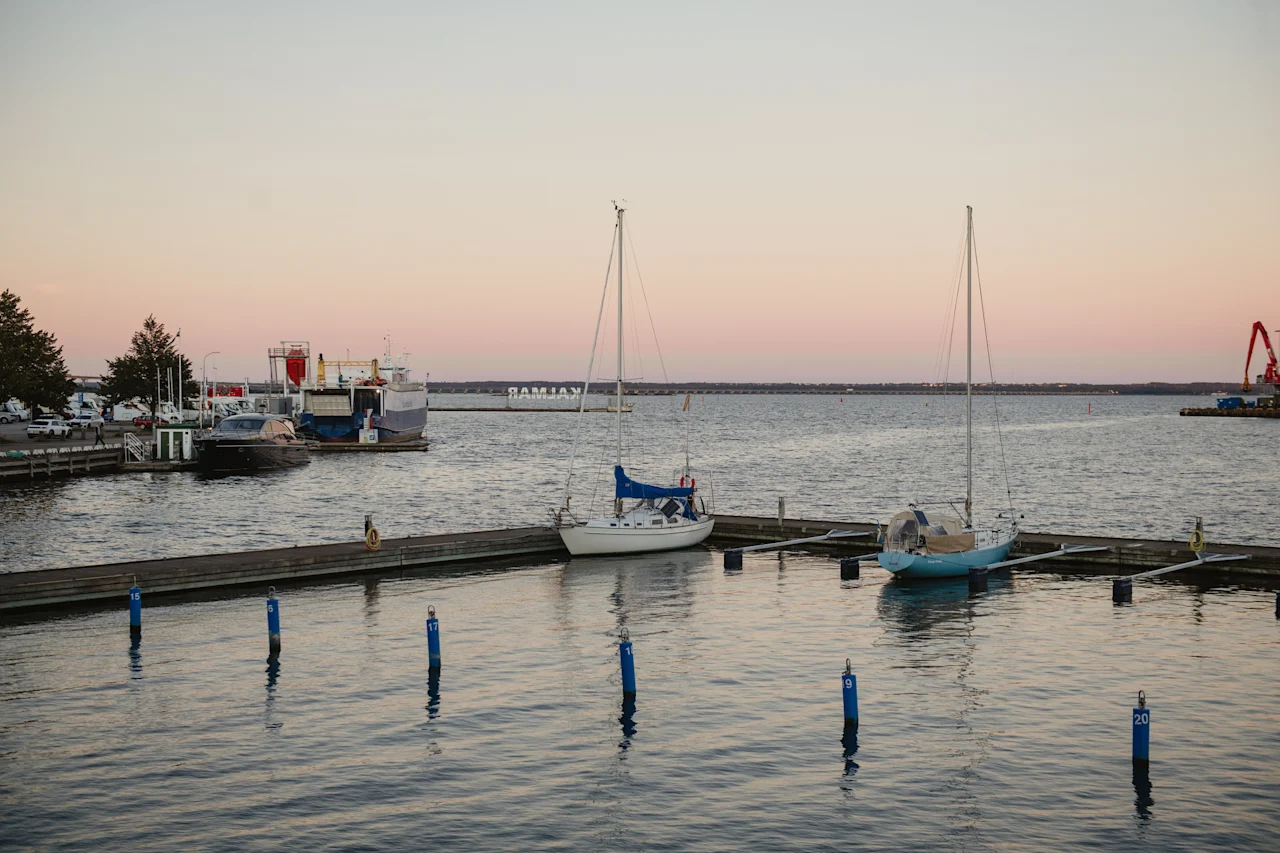 Sejlbåde fortøjet i en rolig marina under en blød solnedgang, med stille vand og en fjern kystlinje.