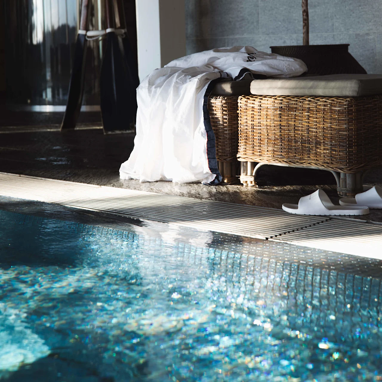 A wicker bench supports a white robe, near a clear blue swimming pool. White slippers rest beside it, with a potted plant and curved metal wall in the background.