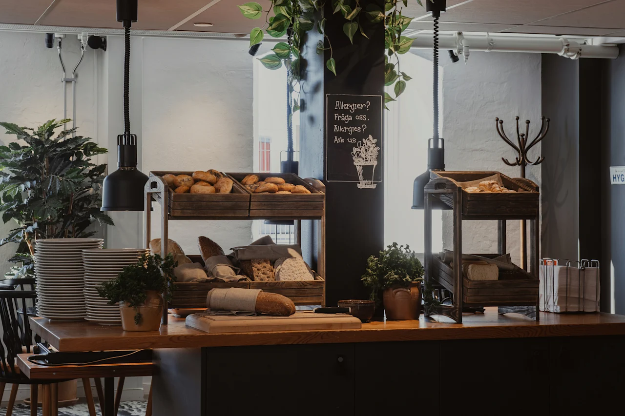Viihtyisä aamiaisbuffet, jossa on erilaisia leipiä ja leivonnaisia puulaatikoissa, pinottuja lautasia ja allergiakyltti.