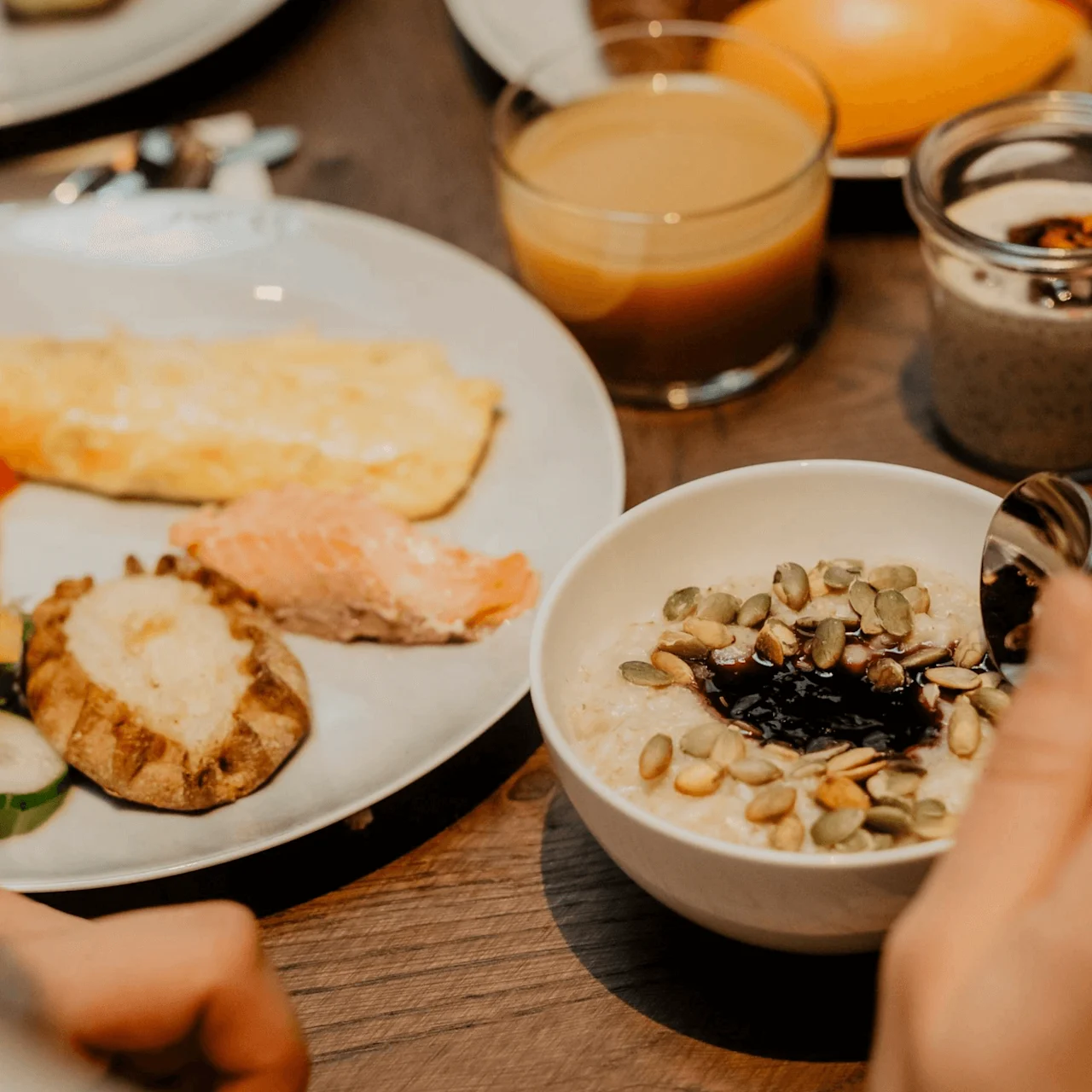 Bowl of oatmeal topped with seeds and jam, held by a person with a spoon; surrounded by plates of cured salmon, vegetables, omelette, juice, and chia pudding on a wooden table.