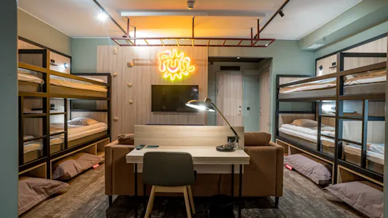 A cozy room features two sets of bunk beds with a sofa and desk in the center. A large TV is mounted on the wall, and a neon "Fun" sign glows above.