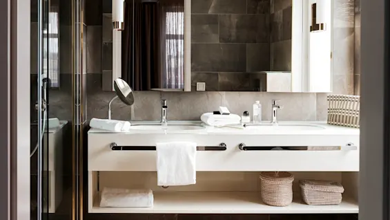 Baderom med en dobbelt vaskebenk med hvite håndklær, toalettartikler og et rundt speil, omgitt av mørke steinfliser. Under står flettede oppbevaringskurver pent plassert.