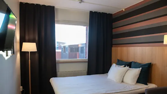 A neatly made bed with white and teal pillows, in a room with striped wallpaper, a floor lamp, a window, and a wall-mounted TV.