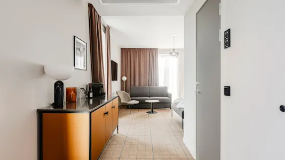 Et moderne hotelværelse med en grå sofa og stol omkring et lille bord. Brune gardiner dækker vinduerne. Et skab har en lampe og kaffemaskine. Neutral indretning.