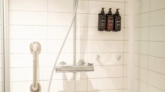 Shower area with three brown bottles on a wall-mounted rack, shiny metal grab bar, and shower fixtures. White tiled walls create a clean, minimalistic bathroom environment.