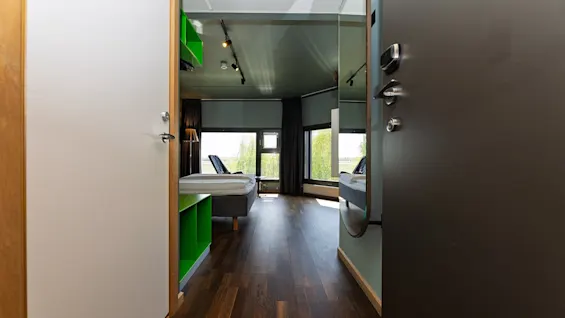 A room with two single beds, chairs, and a window view of greenery, seen through an open doorway with wooden flooring.