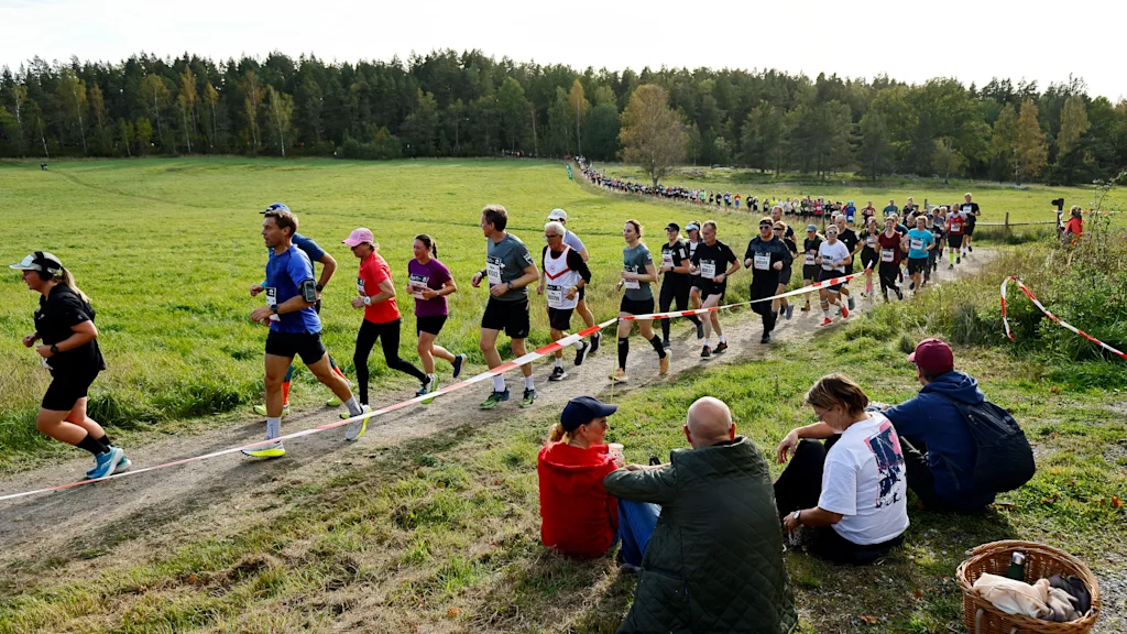 Runners participating in Lidingöloppet on a dirt path through a green field, with spectators watching from the side.