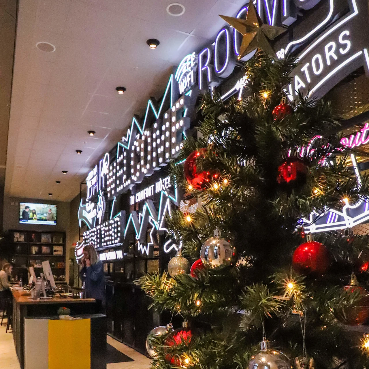 Et dekorert juletre med røde og sølvfargede ornamenter står i en opplyst lounge. Bak er det et neonskilt med teksten "ROOMS". Folk er ved resepsjonsdisken.