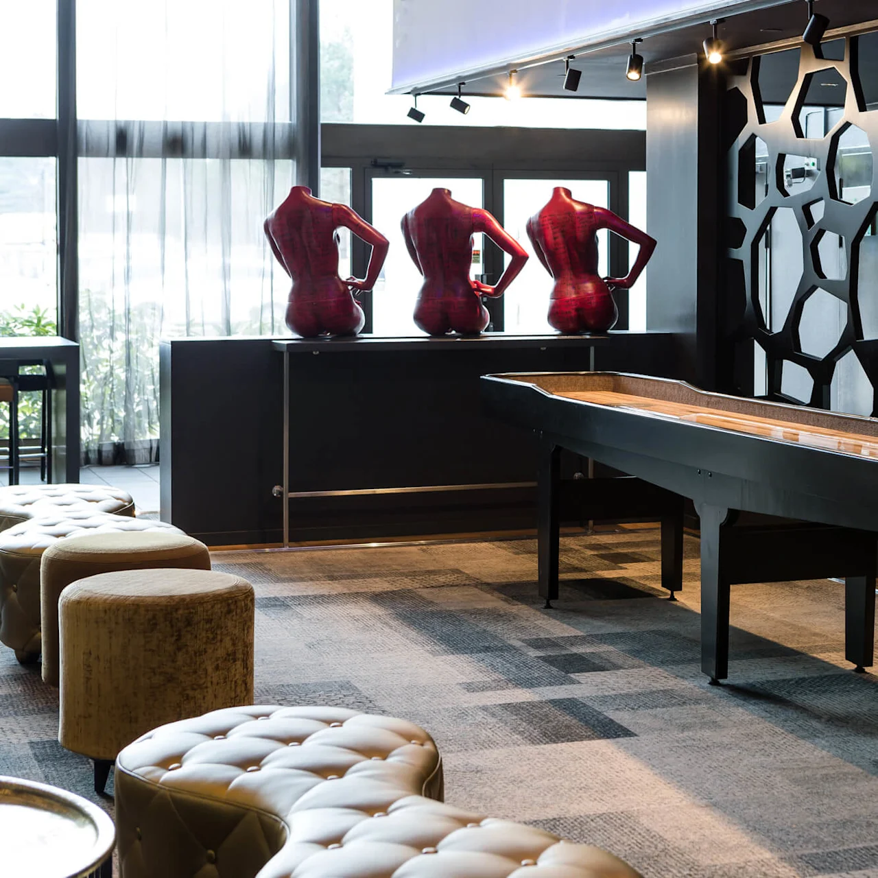 A shuffleboard table stands in a stylish lounge with patterned partitions and mannequin torsos displayed on a counter by large windows. The area features tufted seating and modern decor.