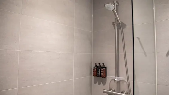 En moderne dusj med lysegrå flislagte vegger, et kromfarget dusjhode og tre mørke flasker med toalettartikler på en dispenser.
