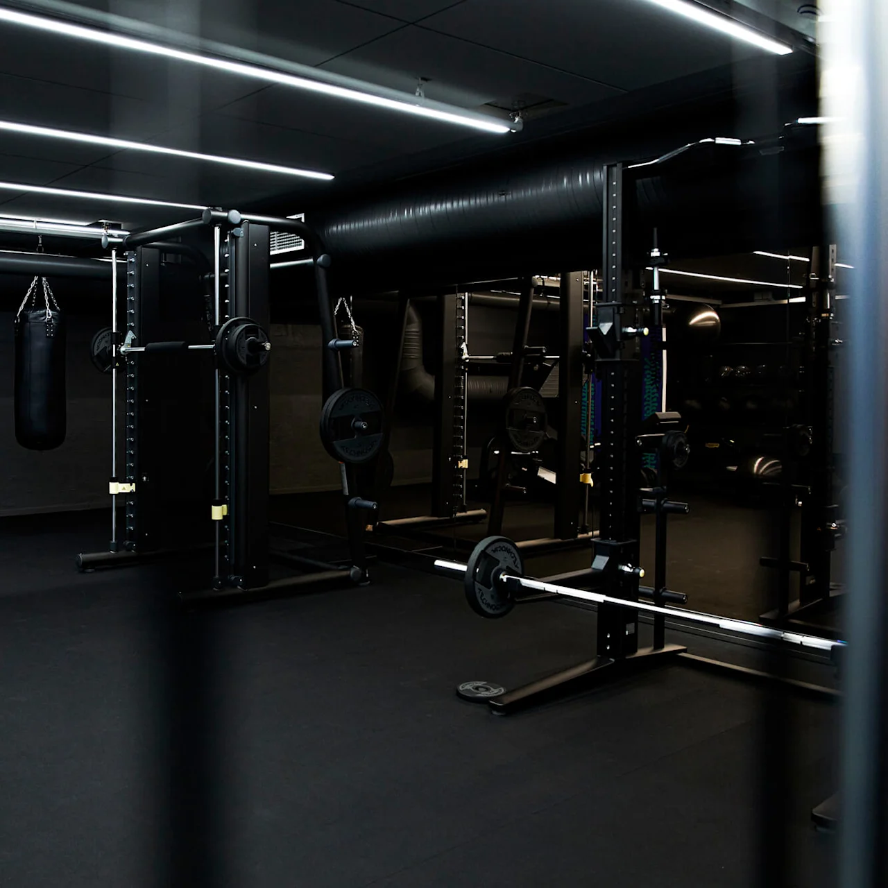 Et fitnesscenter har vægtstativer og maskiner under lyse loftslys, med en boksebold hængende på venstre side, omgivet af et mørkt, glat interiør.