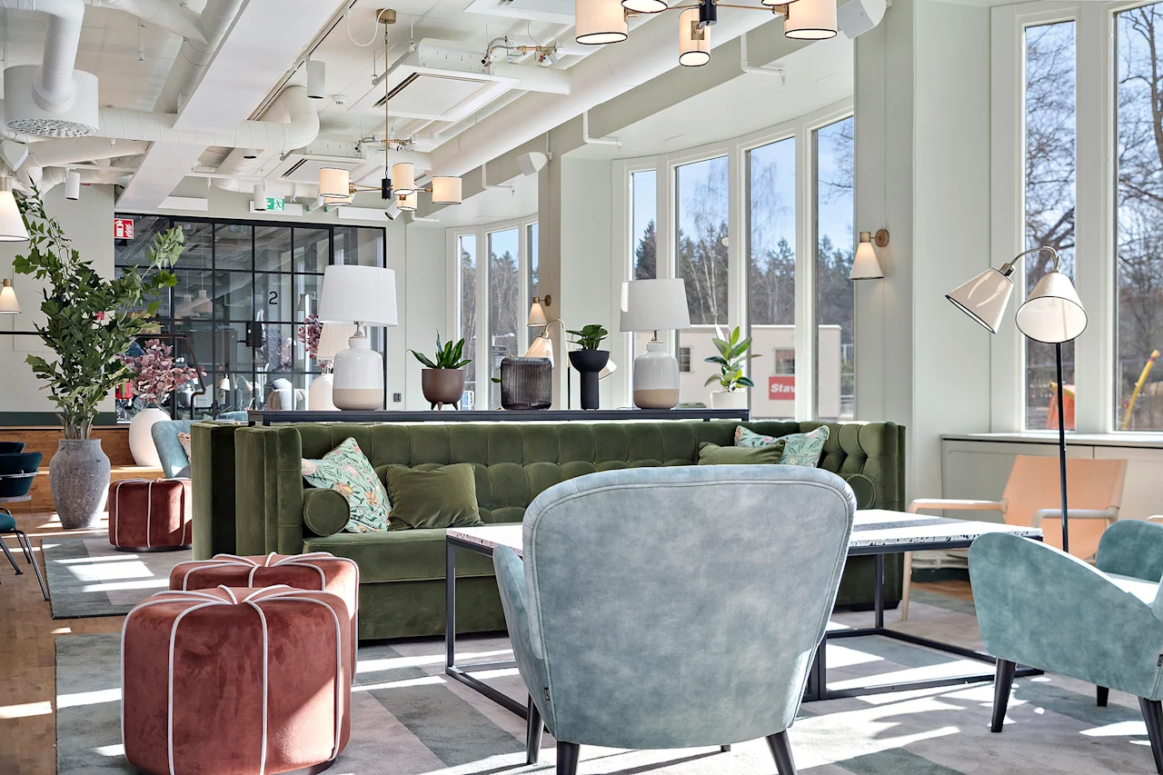Lyst og moderne hotellobbyområde med en grønn fløyelsofa, lenestoler og store vinduer som slipper inn sollys.