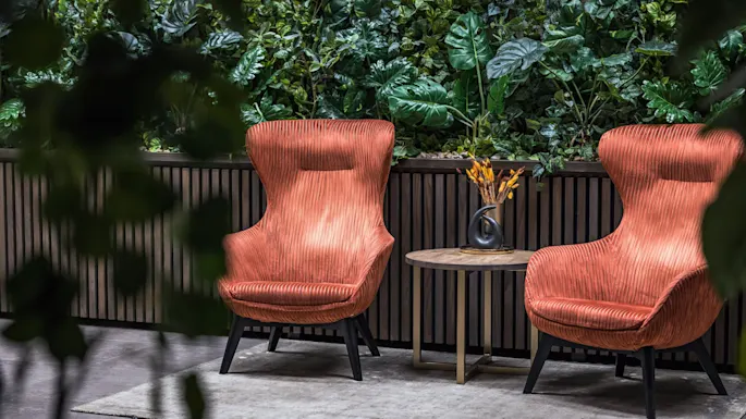 Kaksi oranssia nojatuolia seisovat pienen pyöreän pöydän vieressä mustan maljakon kanssa matolla, jota ympäröivät rehevät vihreät kasvit pystysuoralla puutarhaseinällä.