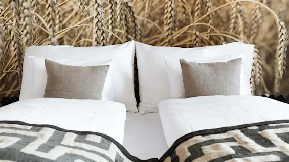 A neatly made bed with white bedding, flanked by wheat sheaves in a rustic setting.