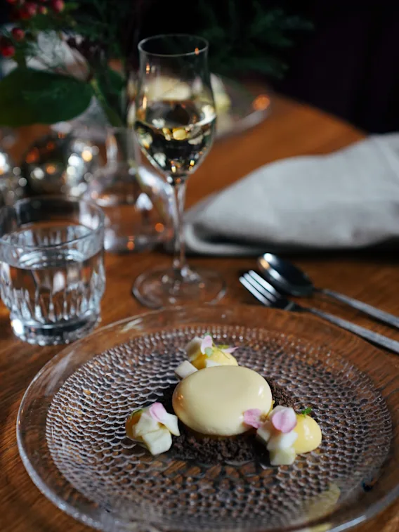 A dessert arranged elegantly on a textured glass plate with garnish, accompanied by a glass of wine and cutlery, set on a wooden table with decorative greenery nearby.