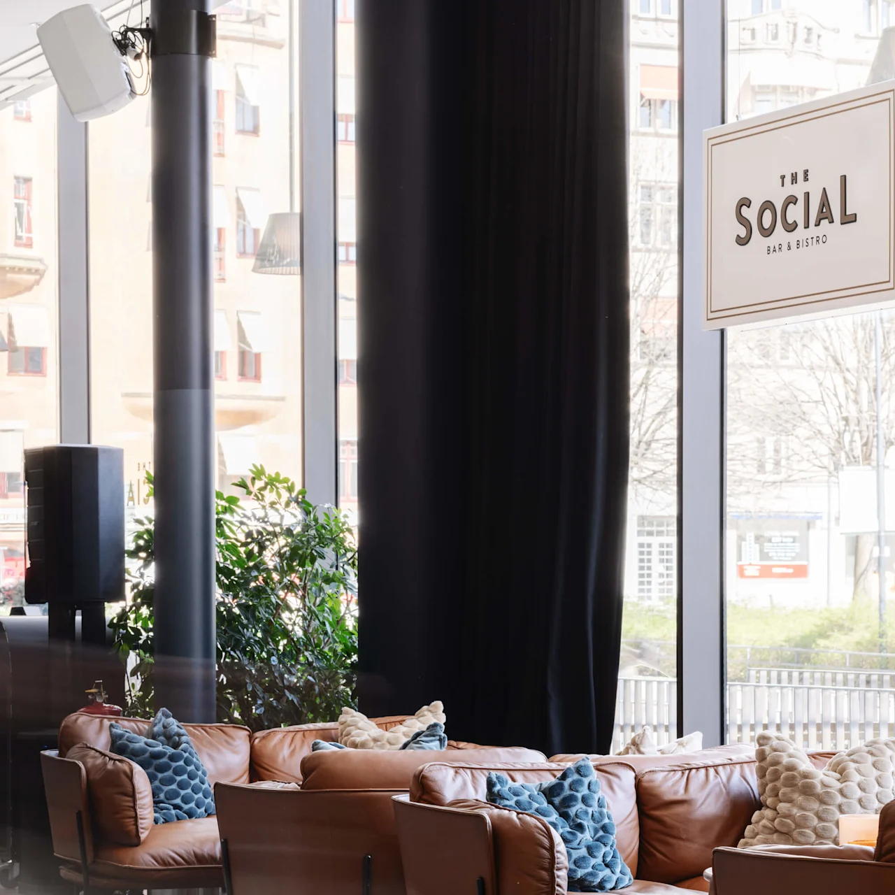 Bruna läderstolar med blå och beige kuddar arrangerade i en sittgrupp nära stora fönster. En skylt lyder "THE SOCIAL BAR & BISTRO."