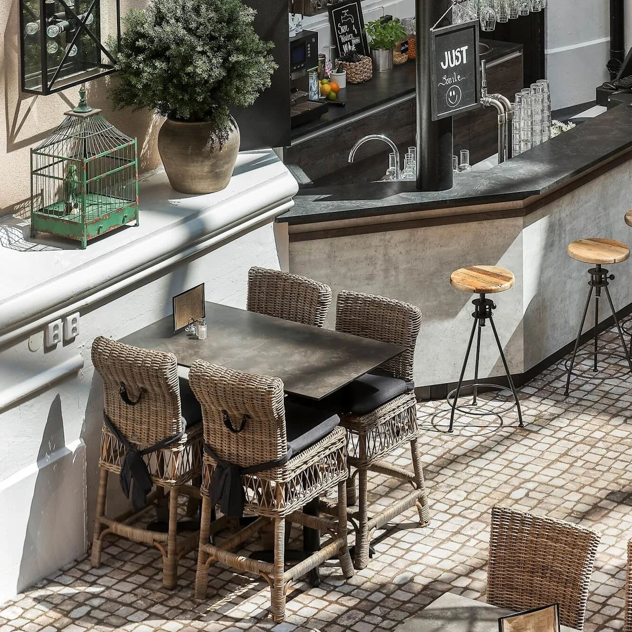 Rottingstolar och bord ordnade i ett soligt utekafé med bardel och en grön fågelbur på kanten.