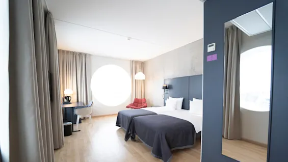 A hotel room with two neatly made twin beds, walls painted dark blue, light wooden flooring, a desk with a lamp, and a large circular window providing natural light.