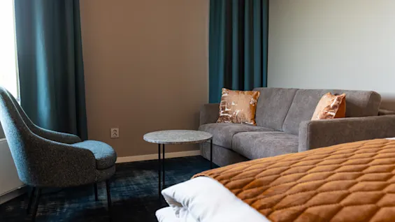 A gray sofa with decorative pillows rests against a wall, facing a round table and armchair. Nearby, a bed with a quilted brown coverlet adds warmth to the cozy room.