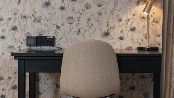 A desk setup with a beige chair, a lit lamp, and floral wallpaper. Power and Ethernet outlets are on the wall.