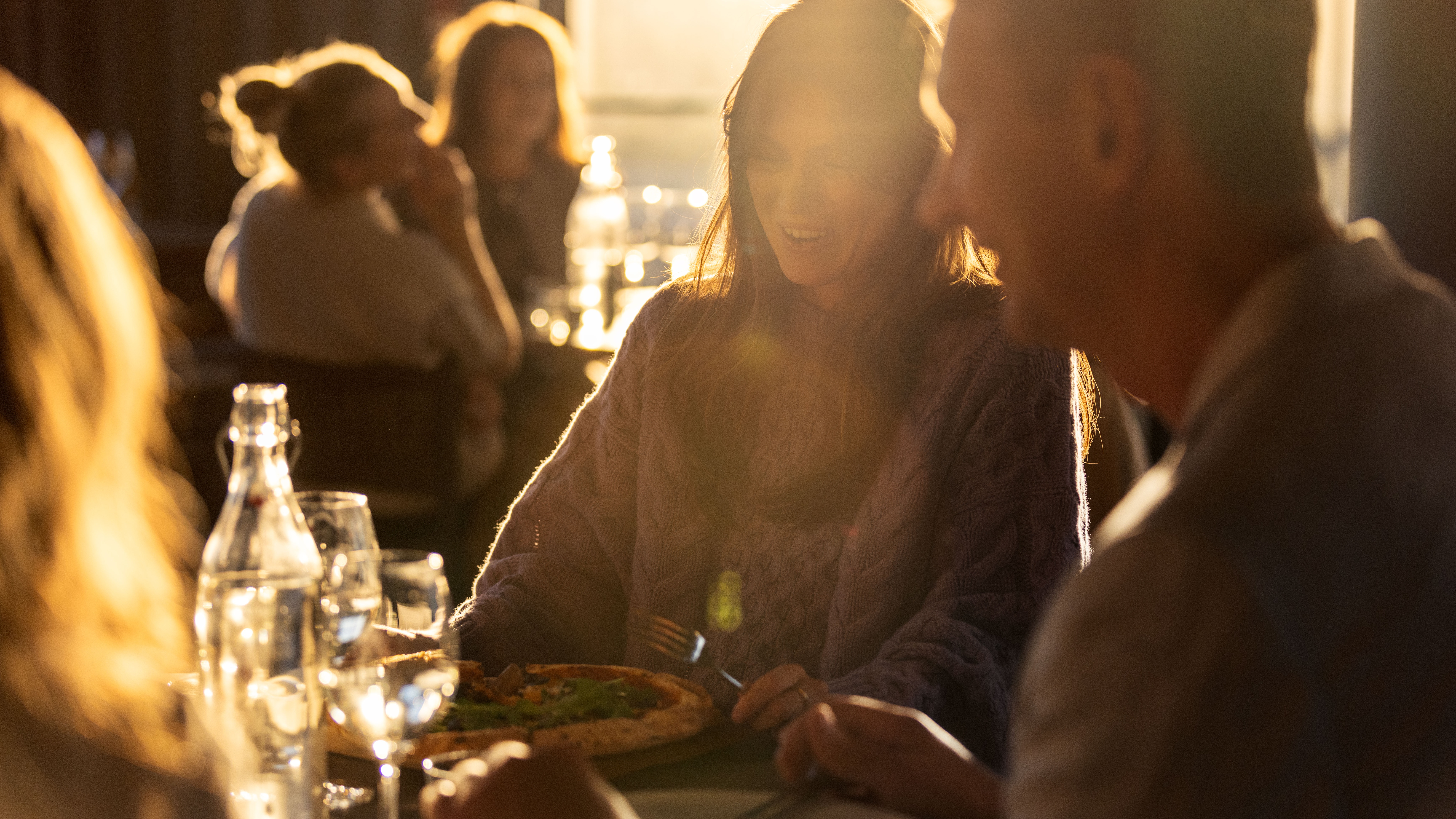 Nainen nauttii pizzasta pöydässä seurassa lämpimästi valaistussa ravintolassa. Auringonvalo suodattuu ikkunasta luoden viihtyisän tunnelman ruokailutilanteessa.